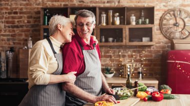 Yaşlı bir çift birlikte yemek hazırlarken mutfakta gülümser ve kucaklaşır. Adam ahşap kesim tahtasıyla sebze kesiyor ve yakınlarda taze sebzeler var..