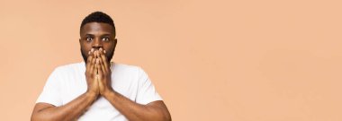 Beyaz tişört giyen ve şeftali rengi arka planda duran sürpriz siyah adam. Elleriyle ağzını kapatıyor ve doğrudan kameraya bakıyor, boşluğu kopyalıyor.