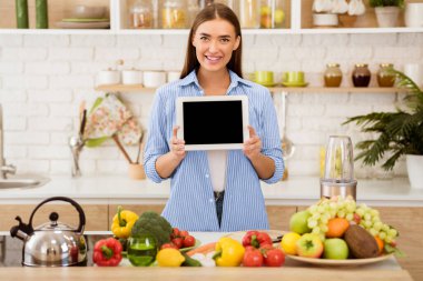 Bir kadın önünde tablet tutarken gülümsüyor, mutfakta taze meyve ve blender ile duruyor..