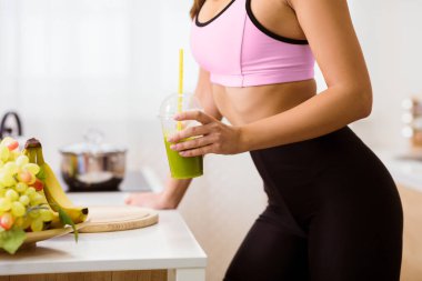 Pembe spor sutyen ve siyah taytlı bir kadın mutfakta elinde yeşil meyve püresi ile dikiliyor. Smoothie 'nin içinde pipet var ve bir içki içmek üzere..