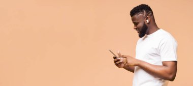 Şeftali rengi arka planda kulaklık takan Afro-Amerikalı bir adam akıllı telefonunu kullanıyor. Telefonuna odaklanmış bir ifadeyle bakıyor, boşluğu kopyala