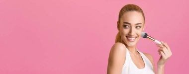 Bu fotoğraf, yanağına makyaj fırçasıyla allık süren parlak bir gülüşü olan genç bir kadını gösteriyor. Beyaz bir kolsuz tişört giyiyor ve saçını at kuyruğu şeklinde yaptırıyor.