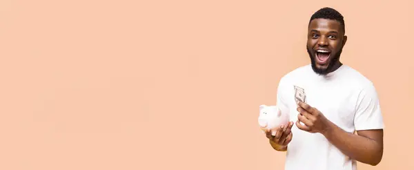 stock image Black man in a white t-shirt is holding cash and a piggy bank in front of a peach-colored background. He is looking at the camera and has a surprised expression on his face, copy space