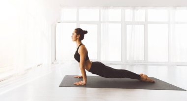 Bir kadın yoga seansında iyi ışıklandırılmış, havadar bir stüdyoda siyah bir paspasın üzerinde yukarı bakan köpek pozu veriyor. Çevre huzuru ve refahı öneriyor.
