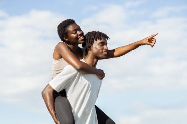 Afro-Amerikalı genç çift güneşli bir günün tadını çıkarıyor. Kadın sırtında erkeğin omuzlarına biniyor ve parmağını gökyüzüne doğrultuyor.