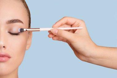 Bu resim, başka bir el tarafından tutulan bir fırçayla göz farını sürerken gözleri kapalı bir kadının yüzünü gösteriyor. Kadın derisi açık, ve resim açık mavi bir arkaplanda..