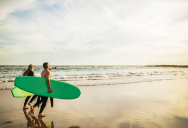 Bir çift sörf yaptıktan bir gün sonra kumlu bir sahilde sörf tahtasıyla yürüyor. Güneş arka planda batıyor ve dalgalar yavaşça kıyıya vuruyor.
