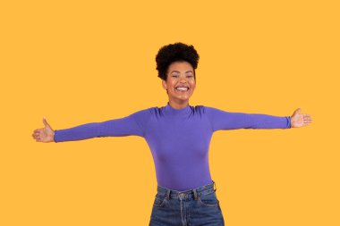 Bu resim kısa, kıvırcık siyah saçlı, mor uzun kollu gömlek giyen ve sarı arka planda duran kot pantolonlu genç bir kadını gösteriyor. Gülümsüyor ve kollarını açıyor.