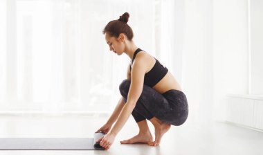 Siyah spor kıyafetleri giymiş genç bir kadın yoga yapıyor, spor paspası katlıyor. Aydınlık, havadar oda onun pratik için sakin ve huzurlu bir ortam sağlar..