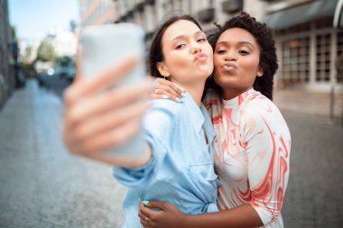 Çok ırklı iki genç kadın, şehir caddesinde selfie çekiyor. İkisi de gülümsüyor ve kameraya bakıyor. Uzun kahverengi saçlı kadın telefonu tutuyor.