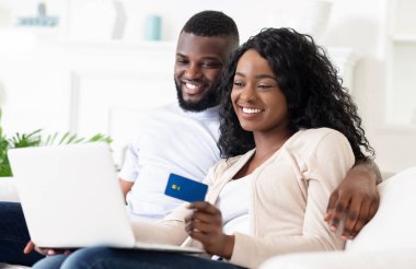 Bir çift kanepede oturuyor, gülümsüyor, online alışveriş için dizüstü bilgisayar kullanıyorlar. Kadının elinde bir kredi kartı var..