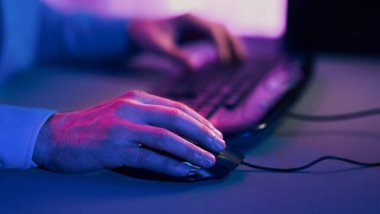 Bir insan eli bilgisayara bağlı olan bir bilgisayar faresinin üzerinde dinlenirken gösterilir. Oda mavi ve mor renklerle aydınlanıyor, dramatik ve renkli bir etki yaratıyor.
