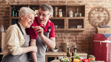 Son sınıf bir çift mutfakta birlikte yemek hazırlarken güler ve gülümser. Adam kadının ağzına doğru bir parça salatalık tutuyor ve kadın da ona gülümsüyor..