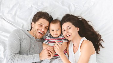 Mutlu bir aile, bir anne, bir baba ve yatakta birlikte yatan küçük bir bebek. Bebekleri onlara bakarken ebeveynler kameraya gülümsüyor. Aile gündelik giysiler giyer.