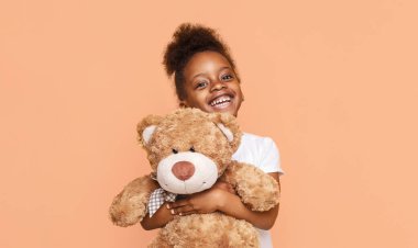Şeftali rengi arka planda kahverengi bir ayıyı tutarken kıvırcık saçlı Afrikalı küçük kız gülümsüyor. O beyaz bir tişört giyiyor ve mutlu görünüyor..