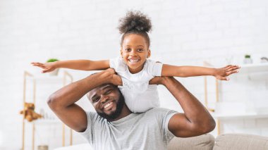 Afrikalı Amerikalı baba ve kızı içeride birlikte oynuyorlar. Baba kızını sırtında taşıyor ve ikisi de gülüyor ve gülüyor..
