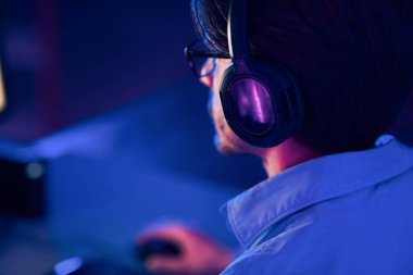 Kulaklık takan bir oyuncu loş bir odada video oyunu oynarken arkadan görülüyor. Oda mavi ışıkla aydınlanıyor, bu da dramatik ve odaklanmış bir atmosfer yaratıyor..