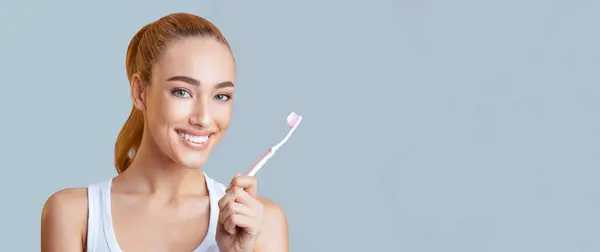 stock image A young woman with red hair and a bright smile is holding a pink toothbrush. She is ready to brush her teeth and keep her oral health in check. She is looking directly at the camera, panorama