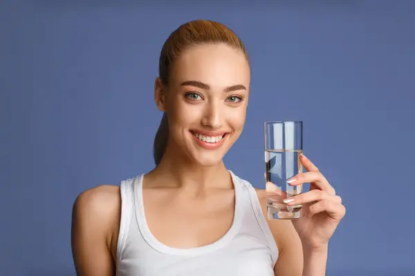 stock image This image shows a young woman with a bright smile holding a glass of water in her right hand. She is wearing a white tank top. The background is a solid blue color.