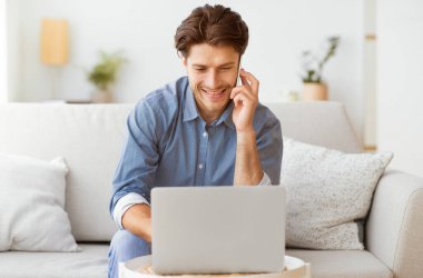 Oturma odasındaki kanepede oturan bir adam dizüstü bilgisayarını kullanıyor ve telefonla konuşuyor. Gülümsüyor ve telefona bakıyor..