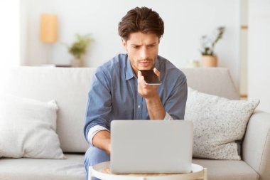 Adamın biri evinde bir kanepede oturuyor ve dizüstü bilgisayar üzerinde çalışırken ses asistanını kullanıyor..