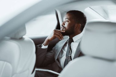 Beyaz tişörtlü, siyah kravatlı, gri takım elbiseli siyah bir adam pencereden dışarı bakan bir arabanın arka koltuğunda oturur, derin düşüncelere dalmıştır. Düşünürken eli çenesinde duruyor..