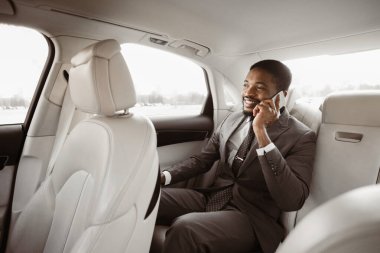 Takım elbiseli siyahi işadamı arabanın arka koltuğunda oturup telefonla konuşuyor. Gülümsüyor ve pencereden dışarı bakıyor. Araba, deri koltuklu lüks bir araca benziyor..