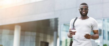 Güneş gözlüklü bir adam elinde kahve fincanı ve elinde bir telefonla gülümsüyor..