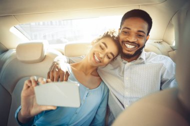 Mutlu bir siyah çift arabanın arka koltuğunda selfie çekiyor. Kadın erkeğin omzuna yaslanırken adam onun elini tutuyor ve gülümsüyor. Görüntü neşe ve samimiyeti yakalar.