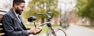 Profesyonel bir siyah adam parkta, tablet ve bisikletle bankta oturuyor. Sahne rahat ve çağdaş, doğal ortamdaki bir üretkenlik anını yakalıyor..