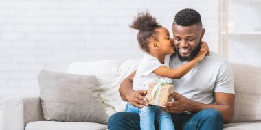 Siyah saçlı Afro-Amerikalı genç kız açık renkli bir kanepede oturuyor ve babasını kucaklarken ona bir hediye veriyor. Baba hediyeye bakarken gülümsüyor..