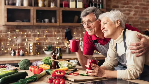 stock image A happy senior couple is enjoying a cup of coffee together in their kitchen. The man is holding a red mug in his hand, while the woman is holding another mug.