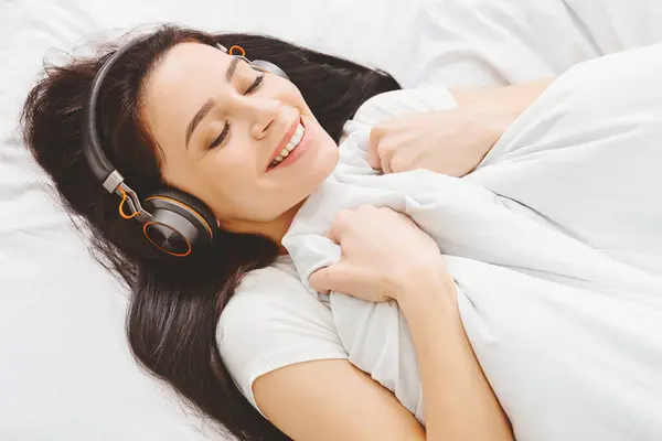 Stock image A woman is laying in bed with headphones on, listening to music. She appears relaxed and immersed in the music playing through the headphones.