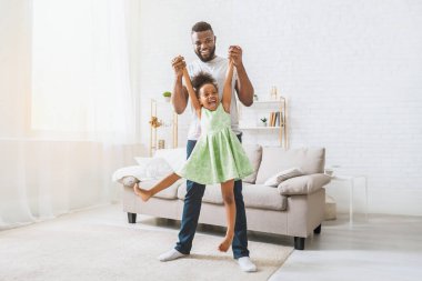 Mutlu bir Afrikalı Amerikalı baba, kızıyla oturma odasında oynuyor. O gülerken ve gülümserken onu ellerinden tutuyor. Her ikisi de gündelik kıyafetler giyiyorlar.
