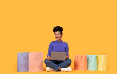 Parlak, dostane bir gülümsemesi olan genç bir kadın sarı bir zeminde dizüstü bilgisayar ve renkli alışveriş torbalarıyla yan yana oturur..