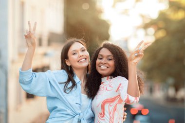 Biri uzun kahverengi saçlı, açık mavi gömlekli, diğeri kıvırcık siyah saçlı ve desenli gömlekli iki genç kadın, şehir ortamında birbirlerine yakın duruyorlar..