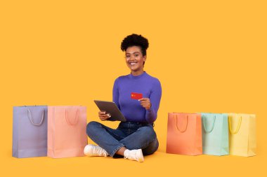 Gülümseyen bir kadın, bir elinde tablet, diğer elinde kredi kartıyla sarı arka planda oturur. Etrafı renkli alışveriş torbalarıyla çevrili..