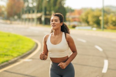 Beyaz spor sutyen ve gri tayt giyen bir kadın banliyö caddesinde koşuyor. Önüne bakıyor ve koşusunun ortasında gibi görünüyor..