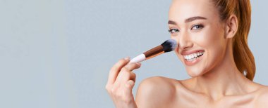 Uzun, sarı saçlı bir kadın fırçayla makyaj yapıyor. Kameraya gülümsüyor ve cildi kusursuz görünüyor. Arkaplan yumuşak mavi bir renk, boşluğu kopyala