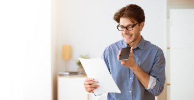 Mavi tişörtlü ve gözlüklü bir adam elinde bir akıllı telefon tutuyor ve bir belgeyi okuyor, sesli mesaj kaydediyor..