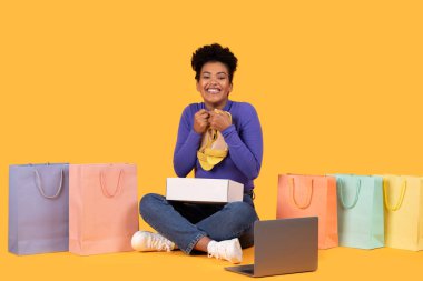 Sarı arka planda oturan genç bir kadın etrafında birkaç alışveriş torbası var. Elinde bir çift yeni ayakkabı tutuyor, heyecanlı ve mutlu görünüyor. Yerde bir laptop var.