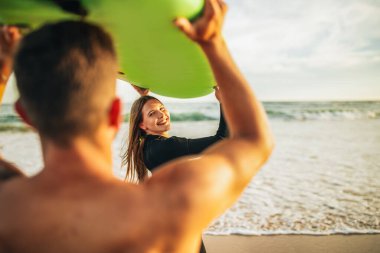 Bir çift, kafalarının üstünde yeşil bir sörf tahtasıyla kumlu bir plajda duruyor. Adam kameradan yüzünü çeviriyor kadın da kameraya bakıp gülümsüyor..