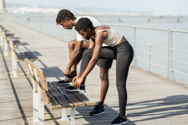 Spor elbiseli siyah çift ayakkabılarını suya bakan ahşap bir bankta bağlıyor. Kadın bağcıklarını bağlamak için eğilirken adam diz çöker..