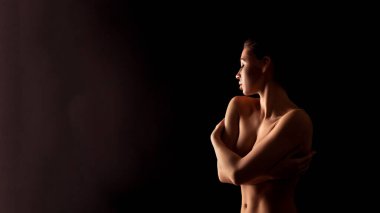 Bu görüntü, karanlık bir odada kolları göğsünde çapraz duran bir kadını gösteriyor. Işıklandırma yumuşak ve vücudunu ve yüzünü silüetle aydınlatıyor..