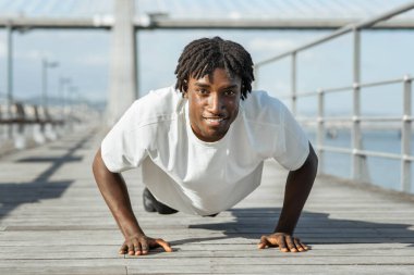 Afrika kökenli, rastalı bir adam, okyanusun kıyısındaki ahşap bir güvertede şınav çekiyor. Beyaz bir tişört giyiyor ve iyi durumda gibi görünüyor. Gülümsüyor ve kameraya bakıyor.