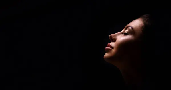 stock image A close up shot of a womans face with eyes closed, lit from the side, with a dark background. She is turned slightly away from the camera. Her lips are closed and she appears to be relaxed.