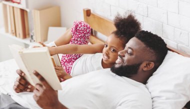 Kara baba ve küçük kızı yatakta uzanıyorlar, baba ona sesli bir kitap okuyor. Kız omzuna yaslanıyor, dikkatlice dinliyor, baba gülümsüyor ve kitaba bakıyor..