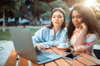 İki genç etnik çeşitlilikte kadın bir kafenin dışında bir masada oturuyor ve bir dizüstü bilgisayar üzerinde çalışıyor. Görüntü, uzaktan çalışan veya bir projede işbirliği yapan insanların ortak bir sahnesini yakalar..