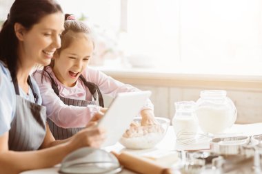 Bir anne ve kızı mutfakta birlikte yemek pişiriyorlar. Bir tablete bakıyorlar, muhtemelen bir tarif için. Masanın üzerinde çeşitli malzemeler ve aletler var, boşluğu kopyala