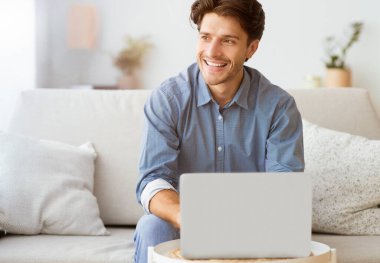 Oturma odasındaki koltukta oturan bir adam yan tarafa bakarken gülümsüyor. Dizüstü bilgisayar kullanıyor ve eğleniyor gibi görünüyor..